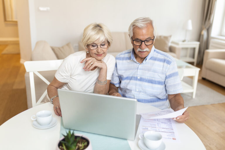Happy middle aged husband and wife sitting at table with laptop and paper bills, calculating domestic incomes together at home.